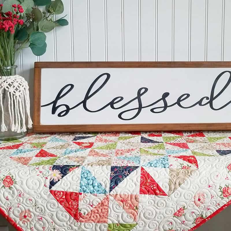 Moda Summer Charm Swap: Charm Square Table Runner by popular quilting blog, A Quilting Life: image of a Moda pattern and Harper's Garden light fabric charm table runner with a white blessed sign and flower vase resting on top.