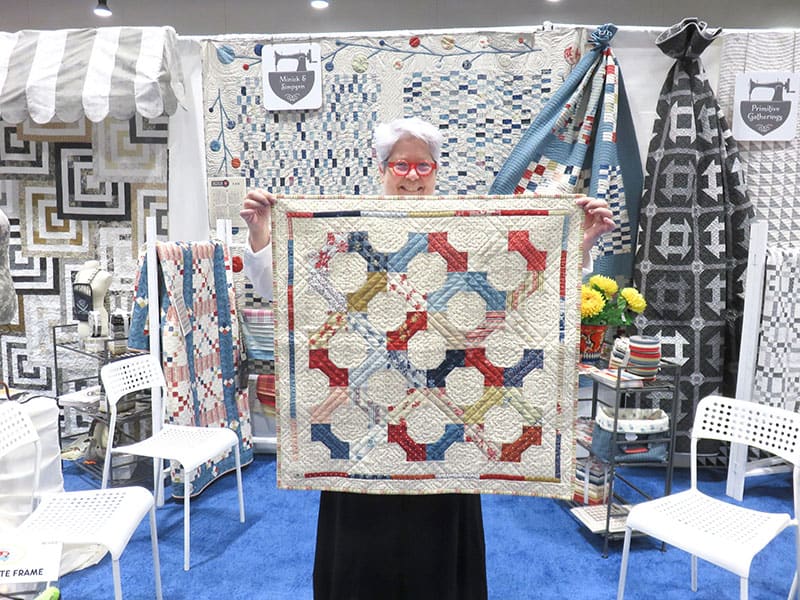 Moda Summer Charm Swap | Table Topper Quilt by popular quilting blog, A Quilting Life: image of a woman holding up a North Port Collection fabric table topper quilt.