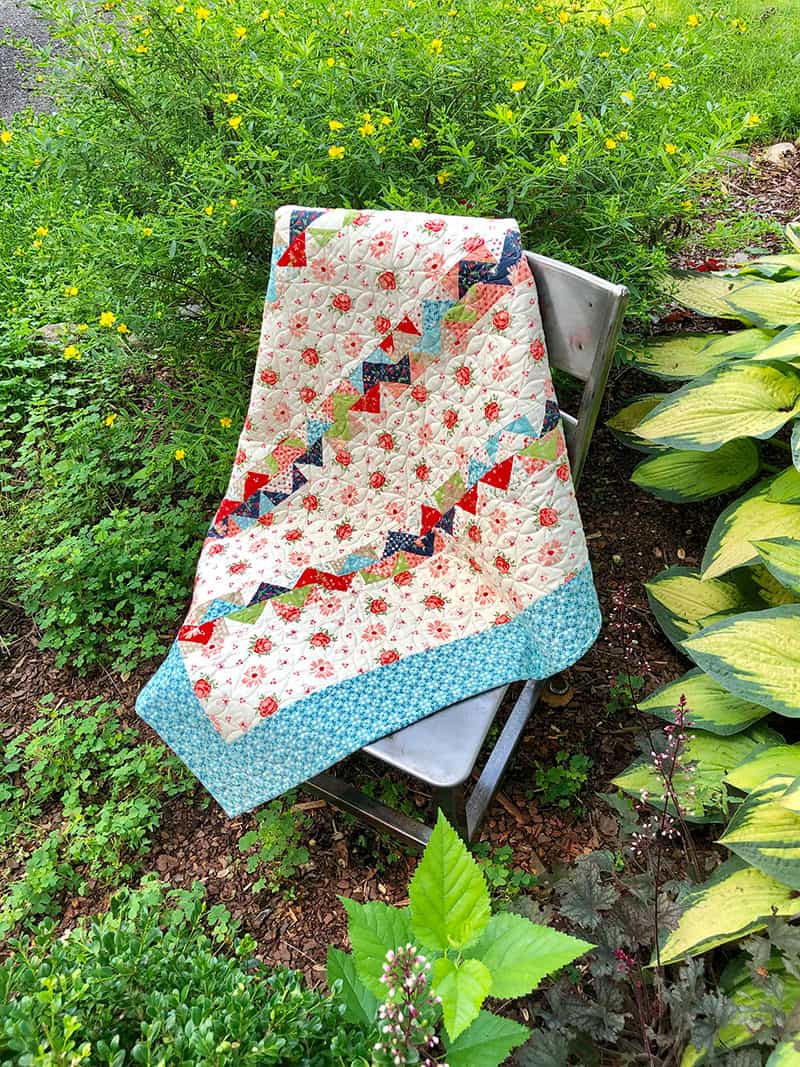 Moda Summer Charm Swap Charm Square Quilt by popular US quilting blog, A Quilting Life: image of a charm square quilt draped over a chair outside in a garden.