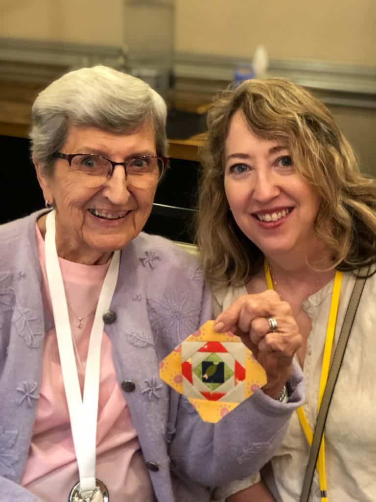 Sherri of a quilting Life | Quiltstock & Restore Quilt Retreats by popular quilting blog, A Quilting Life: image of two women sitting together and holding up a miniature pineapple block made from Sherri and Chelsi fabrics.