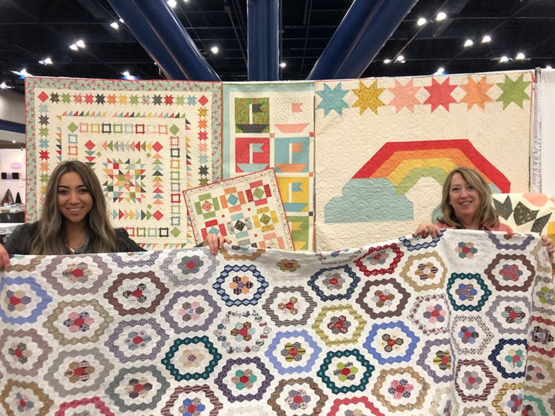 Antique Grandmother's Flower Garden Quilt | International Quilt Market Fall 2019: Part 1 by popular Utah quilting blog, A Quilting Life: image of two women holding up a quilt. 