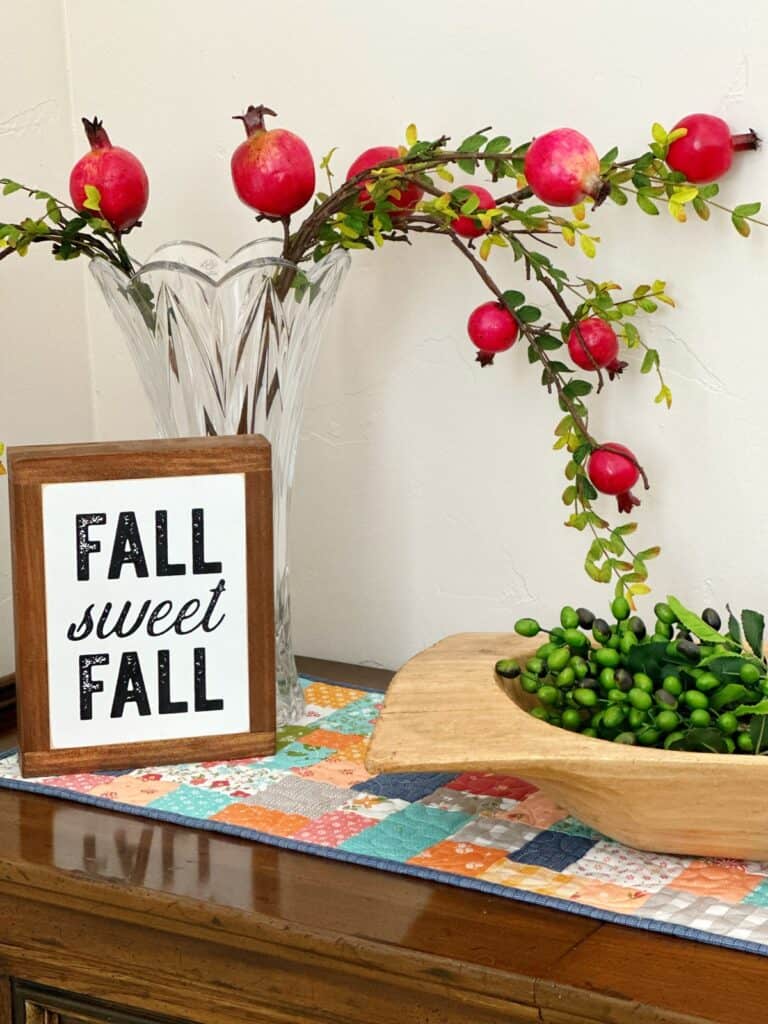 Fall Sweet Fall Sign with wooden bowl, pomegranates and branches on a fall quilted patchwork table runner