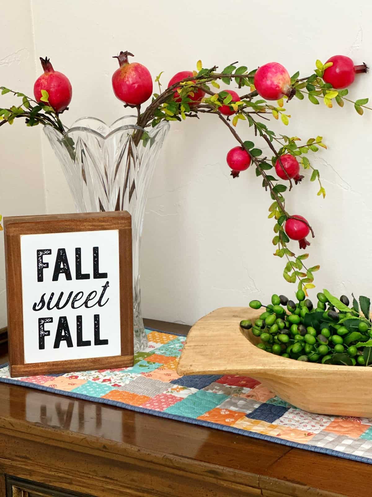 Quilted patchwork table topper with wooden bowl, fall sweet fall sign, pomegranate leaves in vase on side table.