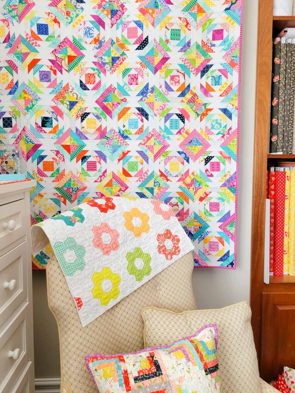 Fabric covered armchair in a corner with pineapple quilt on the wall, log cabin pillow, and hexagon quilted fabric.