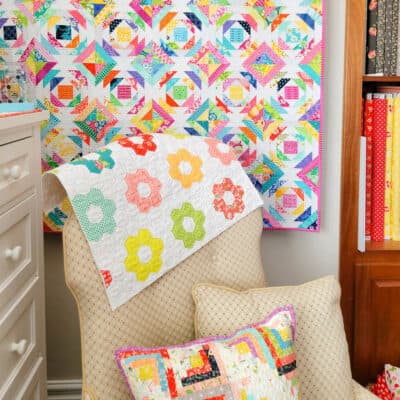 Fabric covered armchair in a corner with pineapple quilt on the wall, log cabin pillow, and hexagon quilted fabric.