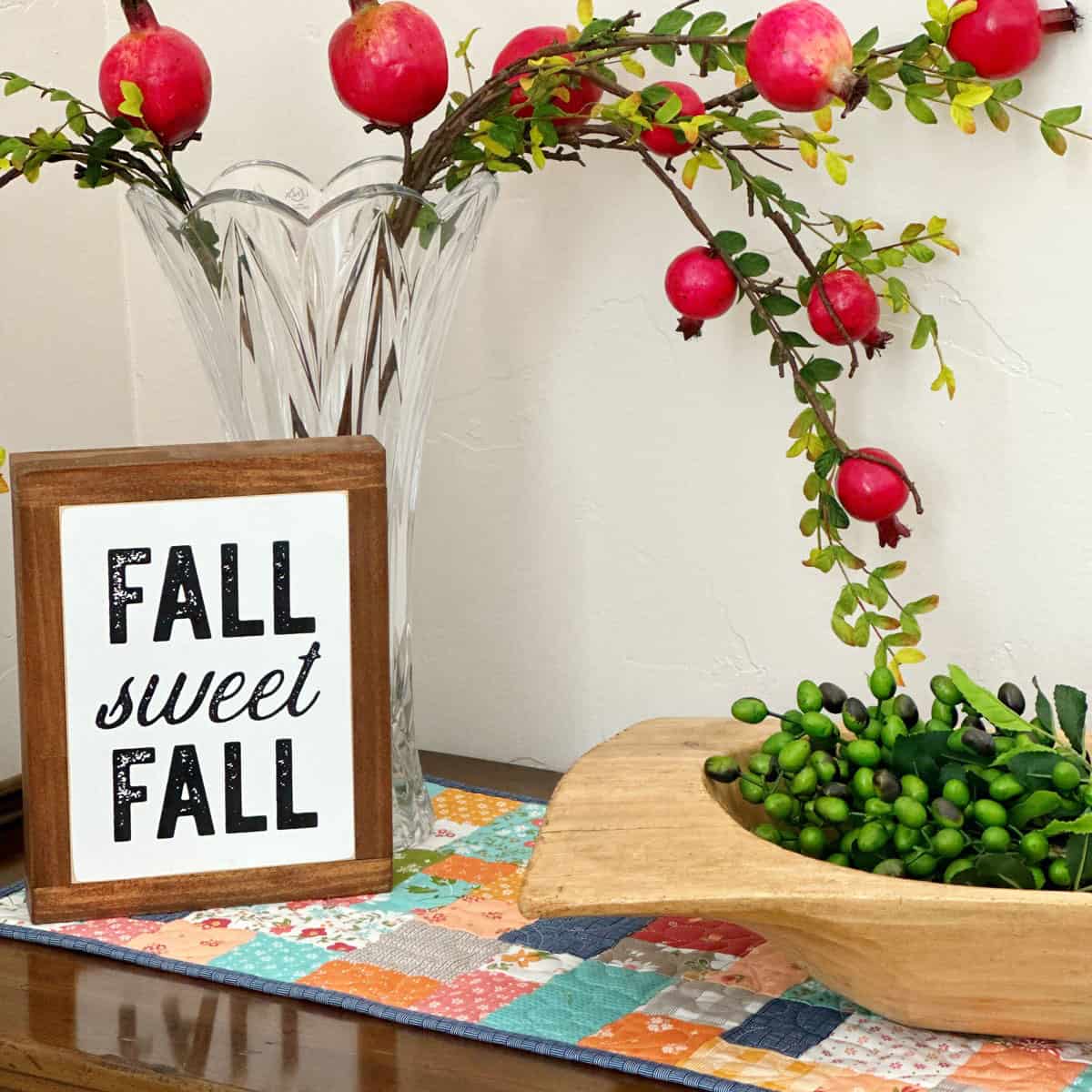 Fall Sweet Fall Sign with wooden bowl, pomegranates and branches on a fall quilted patchwork table runner