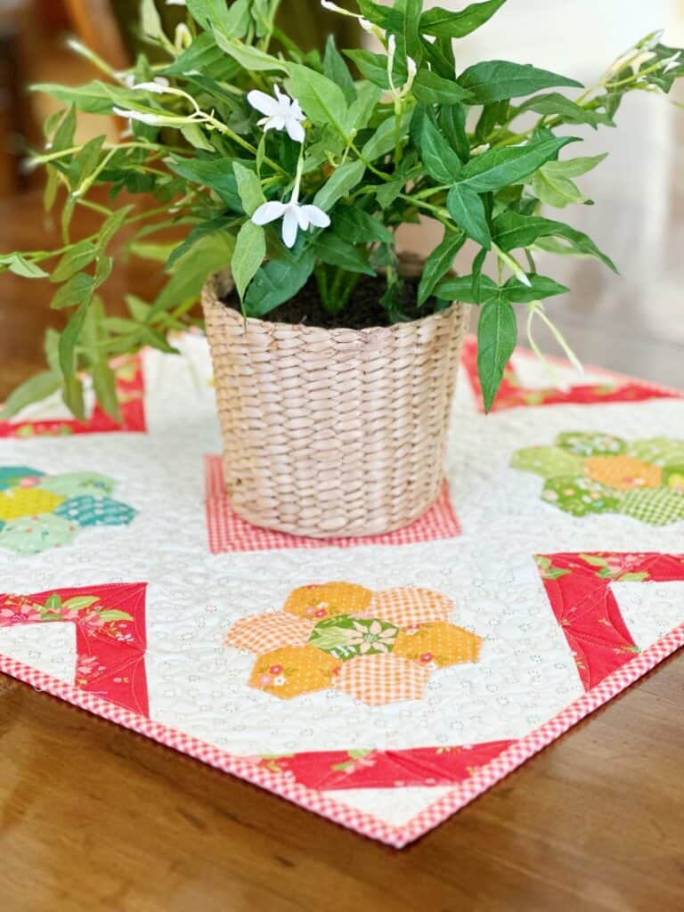 Table topper featuring Grandmother's Flower Garden blocks in bright colors with green plant in basket on top.