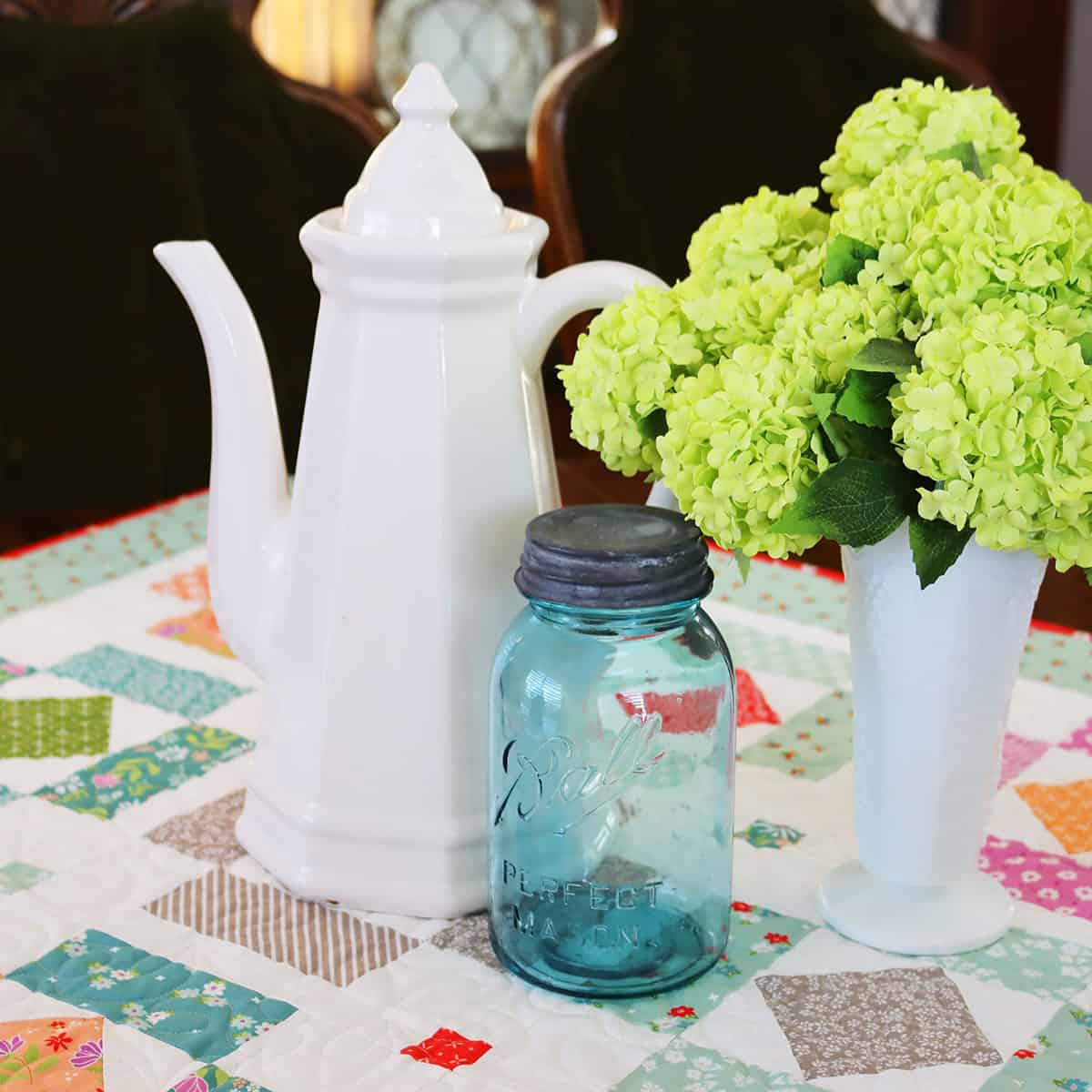 Pitcher, aqua canning jar, and vase with hydrangeas on a quilted table topper.