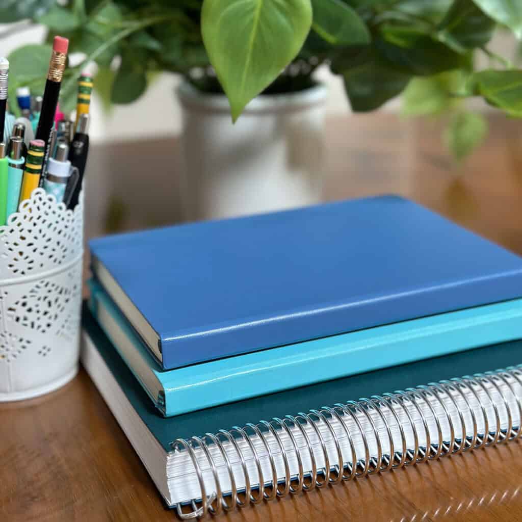 Stack of notebooks on a table with a plant and can of pens and pencils