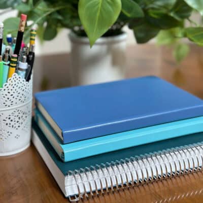 Stack of notebooks on a table with a plant and can of pens and pencils