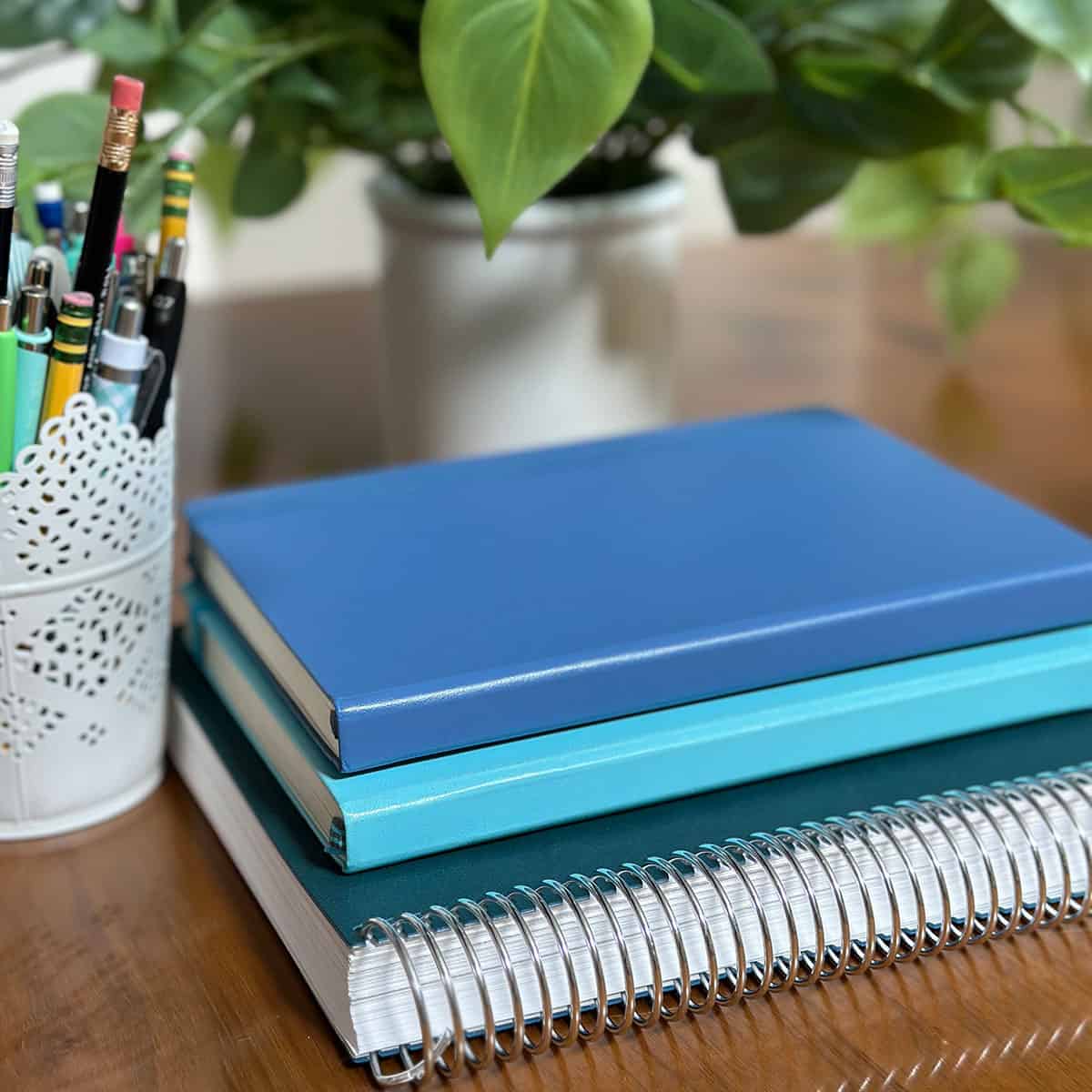 Stack of notebooks on a table with a plant and can of pens and pencils