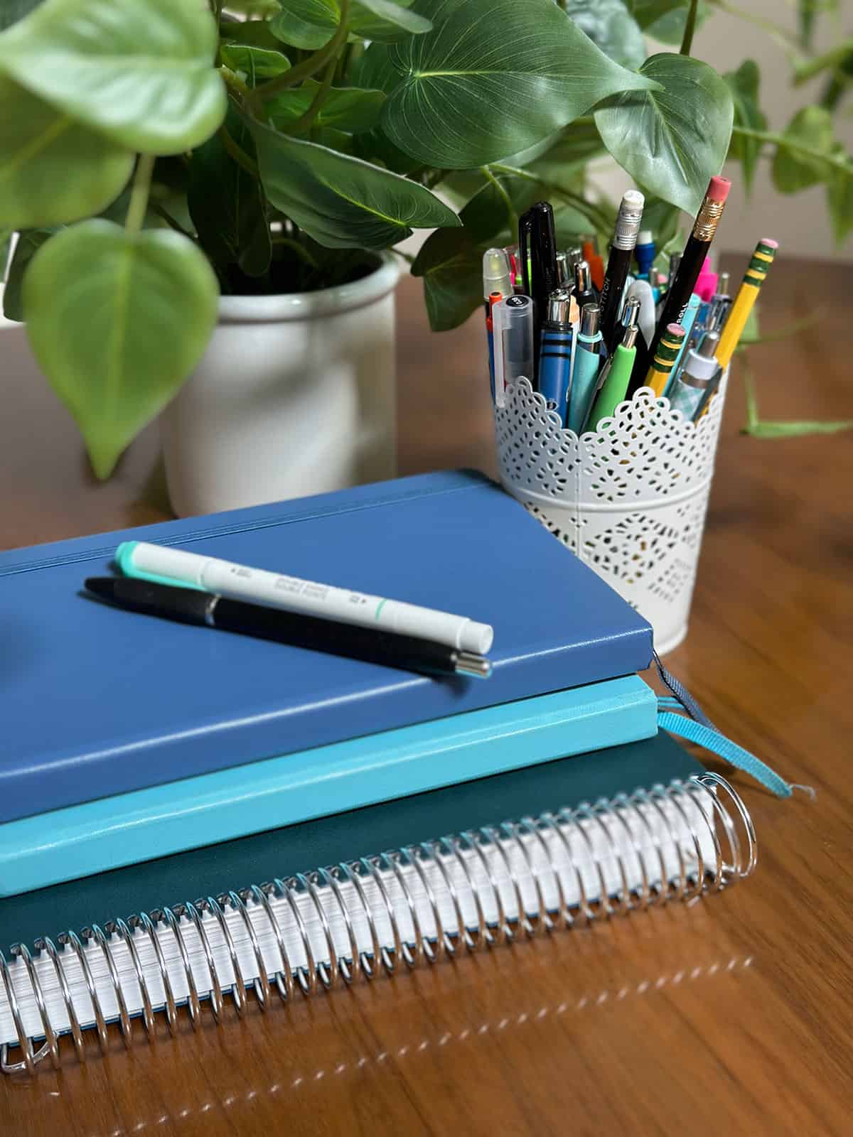 Stack of notebooks on a table with a plant and can of pens and pencils